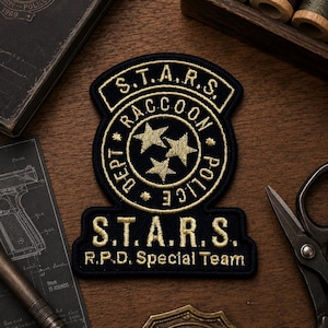 May include: A black and gold embroidered patch featuring the text "S.T.A.R.S. Raccoon Police Dept." and "R.P.D. Special Team." The patch has a shield shape with two gold stars inside a circle. The patch is on a wooden surface.