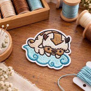 May include: An embroidered patch featuring a stylised cream and brown creature with a dark brown arrow marking on its head, resting on a light blue cloud. The patch has a white border and is crafted with detailed stitching throughout the design.