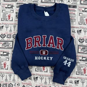 May include: A navy blue crewneck sweatshirt featuring the embroidered text BRIAR U HOCKEY in maroon and white on the chest. The right sleeve displays the embroidered name GRAHAM and the number 44 in white thread. The garment has ribbed cuffs.