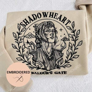 May include: A beige sweatshirt with black embroidered text that reads "Shadowheart Baldur's Gate". The embroidery features a woman with long hair, wearing a dark outfit and a necklace. The design is surrounded by a wreath of leaves.