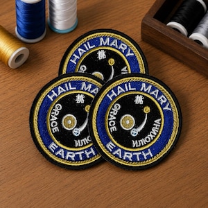 May include: Three circular embroidered patches featuring a black center with a gold and white graphic, surrounded by a blue border with gold trim. The text HAIL MARY and EARTH is embroidered in white, with additional text and symbols in the center.