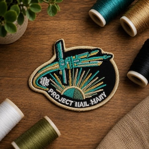 Op de afbeelding: Een geborduurde patch met een zwarte achtergrond, voorzien van een gestileerd ruimteschip in blauwgroen en goud, een zonnestraalpatroon en de tekst PROJECT HAIL MARY in witte hoofdletters, omrand door een gouden stiksel.