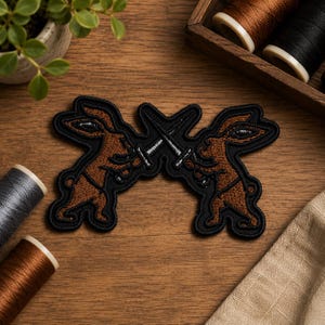 May include: An embroidered patch featuring two brown rabbits standing on their hind legs, each holding a silver sword crossed in the center. The design is outlined in thick black thread, creating a cohesive shape with a dark border against the wood.