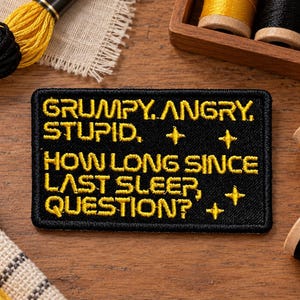 Könnte beinhalten: Ein rechteckiger, schwarz bestickter Aufnäher mit gelbem Text, der lautet: GRUMPY, ANGRY, STUPID. HOW LONG SINCE LAST SLEEP, QUESTION? Der Aufnäher enthält vier kleine gelbe vierzackige Sternsymbole, die zwischen den Textzeilen platziert sind.