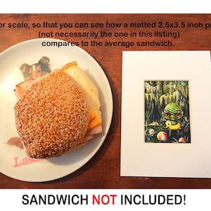 May include: A white matted print of a green creature holding a yellow float in a swamp. The print is 2.5 by 3.5 inches. A sandwich on a plate is shown for scale. The text "SANDWICH NOT INCLUDED!" is printed below the image.