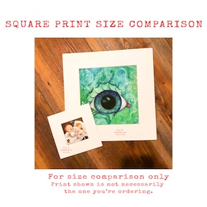 May include: Two square prints of artwork, one with a blue eye and the other with a cartoon illustration. The larger print is matted to 12x12 inches and the smaller print is matted to 6x6 inches. The text "For size comparison only. Print shown is not necessarily the one you're ordering." is below the prints.