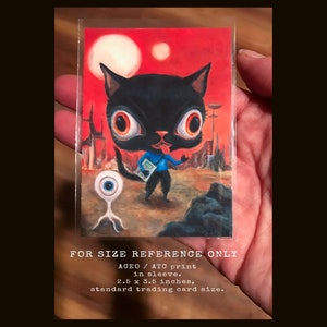 May include: A black cat with large eyes and a blue shirt stands on a red and brown landscape with a single eye creature in the foreground. The image is a 2.5 x 3.5 inch ATC print in a sleeve. FOR SIZE REFERENCE ONLY.