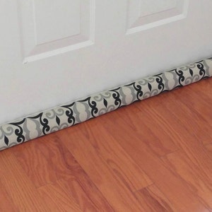 May include: A beige and black patterned doorstop with a cylindrical shape. The doorstop is placed on a wooden floor.