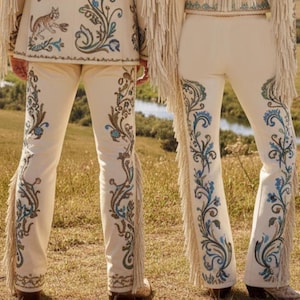 May include: Cream-colored pants with intricate embroidery in shades of blue, brown, and green. The pants feature a flared leg design and fringe detailing along the sides. The ensemble includes a matching jacket with similar embroidery and a cat design.