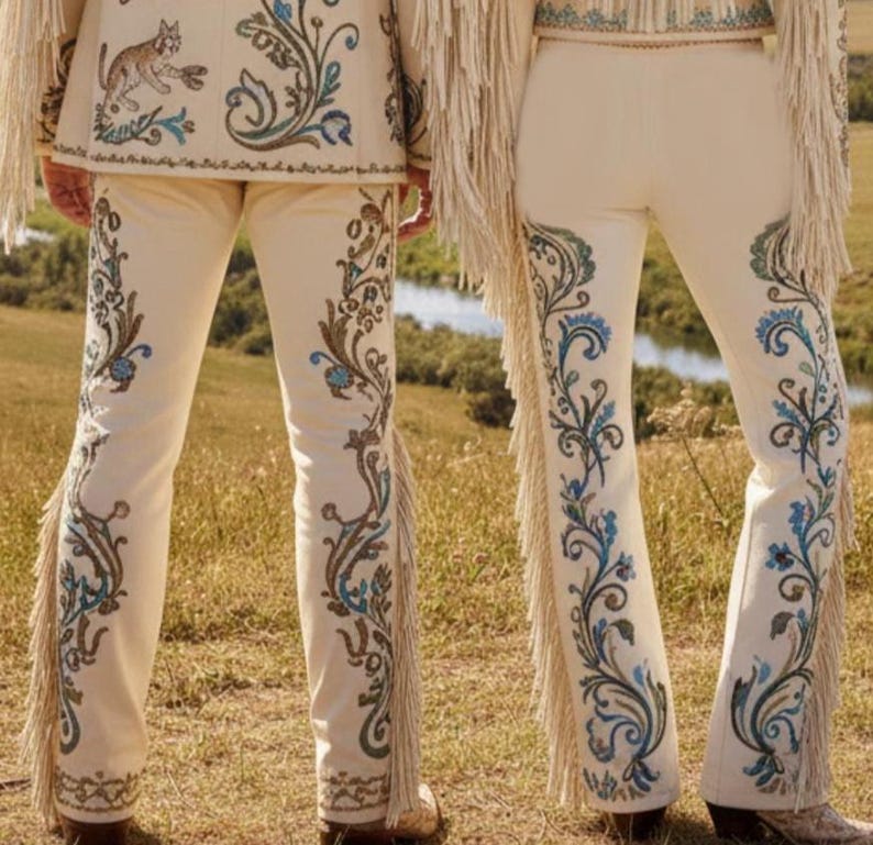 May include: Cream-colored pants with intricate embroidery in shades of blue, brown, and green. The pants feature a flared leg design and fringe detailing along the sides. The ensemble includes a matching jacket with similar embroidery and a cat design.
