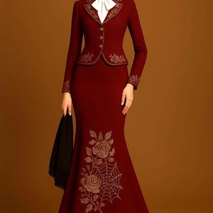 Puede incluir: Un traje de falda burdeos con bordados florales dorados. El traje incluye una chaqueta entallada con una camisa de cuello blanco y una falda larga de estilo sirena. Un chal negro se sostiene en la mano izquierda.