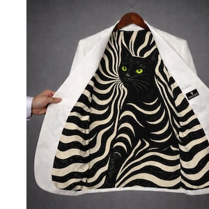 Op de afbeelding: Een witte blazer met een zwart-witte voering met een zwarte kat met groene ogen. De blazer hangt aan een houten hanger. De voering heeft een wervelend patroon.