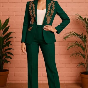 Puede incluir: Un traje pantal&oacute;n verde oscuro con bordados dorados en la chaqueta. El traje incluye una chaqueta entallada con solapas y pantalones rectos a juego. La modelo lleva un top blanco. El fondo es una pared de ladrillo rojo claro.