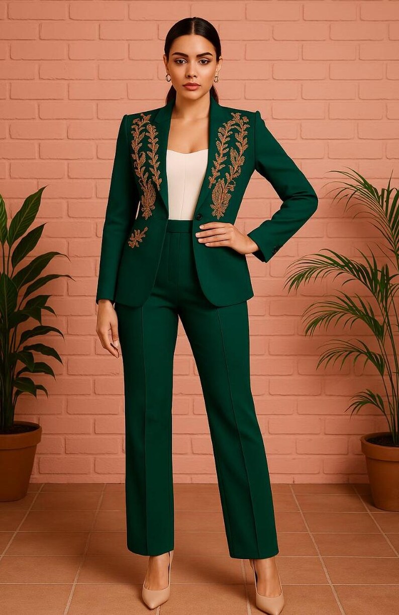 Puede incluir: Un traje pantal&oacute;n verde oscuro con bordados dorados en la chaqueta. El traje incluye una chaqueta entallada con solapas y pantalones rectos a juego. La modelo lleva un top blanco. El fondo es una pared de ladrillo rojo claro.