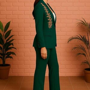 Puede incluir: Un traje pantal&oacute;n verde oscuro con una americana entallada con bordados dorados en la solapa. El traje se combina con pantalones a juego y tacones nude. El fondo incluye una pared de ladrillo y plantas en macetas.