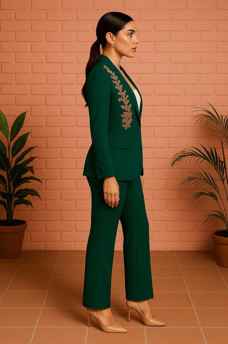 Puede incluir: Un traje pantal&oacute;n verde oscuro con una americana entallada con bordados dorados en la solapa. El traje se combina con pantalones a juego y tacones nude. El fondo incluye una pared de ladrillo y plantas en macetas.