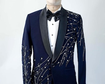 Puede incluir: Una chaqueta de esmoquin azul marino con solapa de satén negro y pajarita negra. La chaqueta presenta una camisa blanca y está adornada con elementos decorativos plateados y blancos. El texto "MAGNATE ARTISAN BESPOKE STUDIO" es visible.