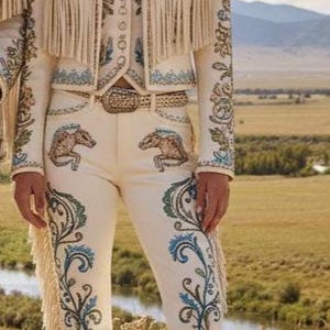 May include: Cream-colored Western-style outfit with intricate embroidery and fringe detailing. The ensemble includes a jacket, vest, and pants, complemented by a cowboy hat. The pants feature horse designs. The outfit is set against a backdrop of a natural landscape.