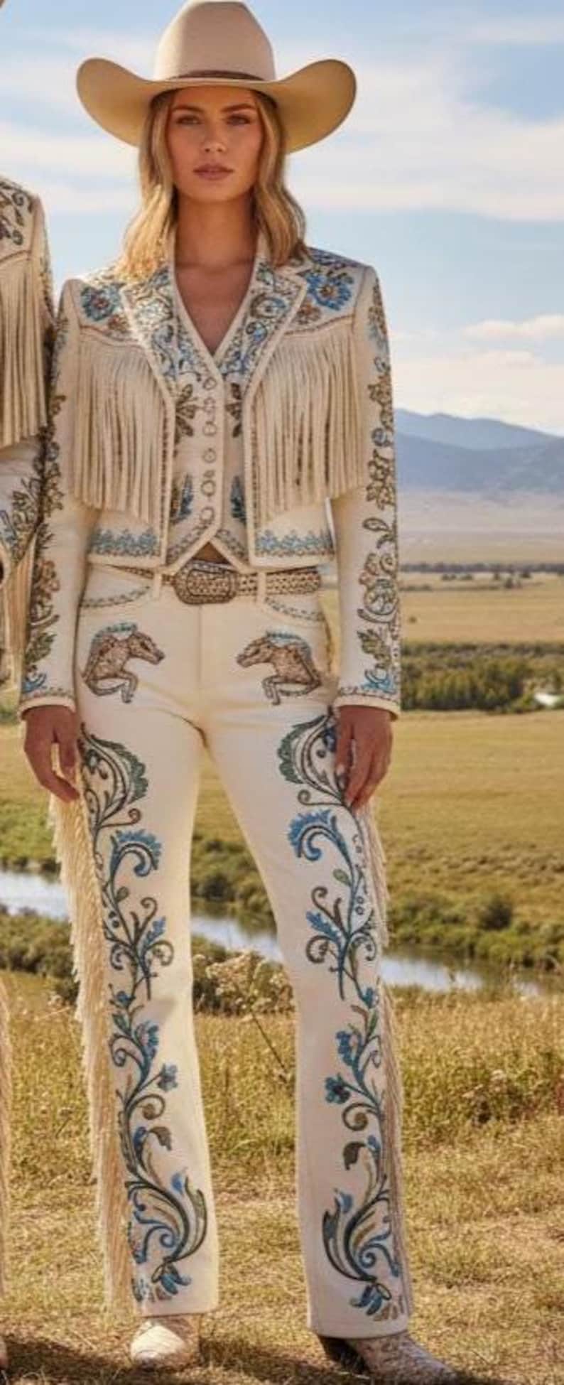 May include: Cream-colored Western-style outfit with intricate embroidery and fringe detailing. The ensemble includes a jacket, vest, and pants, complemented by a cowboy hat. The pants feature horse designs. The outfit is set against a backdrop of a natural landscape.