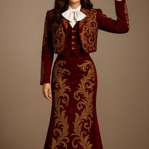 Puede incluir: Un atuendo de mariachi burdeos con bordados dorados. El conjunto incluye una chaqueta, un chaleco y una falda, todos adornados con patrones dorados ornamentados. Un sombrero a juego con detalles dorados y una pajarita blanca completan el look. El atuendo es de estilo tradicional.