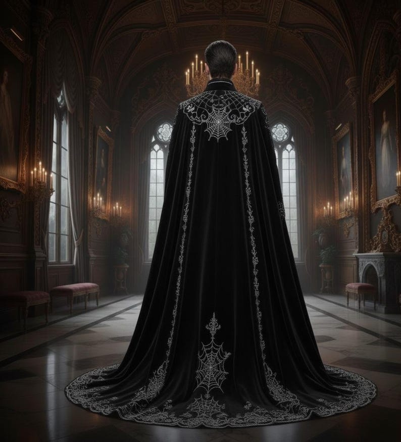 Op de afbeelding: Een lange, zwarte fluwelen cape met ingewikkelde zilveren spinnenweb- en bloemenborduurwerk. De cape heeft een hoge kraag en een lange sleep, perfect voor gothic- of fantasie-evenementen. De afbeelding is geplaatst in een grote, met kaarsen verlichte zaal.