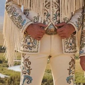 May include: Cream-colored cowboy suit with intricate embroidery in teal and brown, featuring fringe on the jacket and pants. The outfit includes a matching vest, a bolo tie with a turquoise stone, and a cowboy hat.