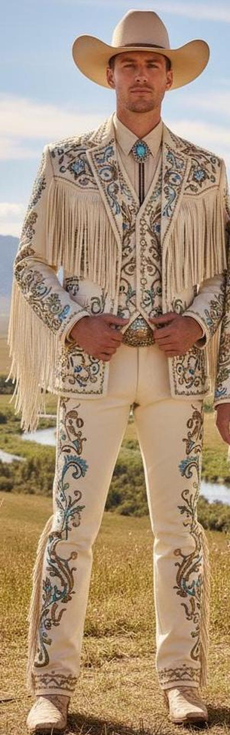 May include: Cream-colored cowboy suit with intricate embroidery in teal and brown, featuring fringe on the jacket and pants. The outfit includes a matching vest, a bolo tie with a turquoise stone, and a cowboy hat.