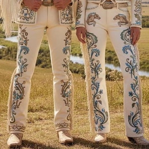 May include: Cream-colored, embroidered pants with intricate floral and scroll designs in blue, green, and brown. The pants feature fringe along the outer seams and are paired with matching boots. The image showcases a traditional Western wear style.
