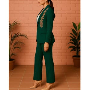 Puede incluir: Un traje pantal&oacute;n verde oscuro con bordados dorados en la solapa de la blazer. El traje se combina con tacones color nude. El fondo presenta una pared de ladrillo y plantas en macetas, creando un look sofisticado y elegante.