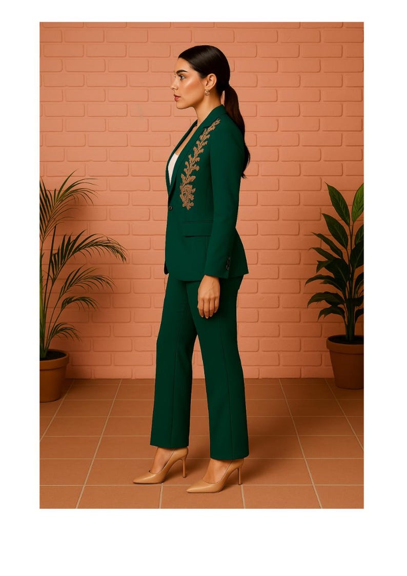 Puede incluir: Un traje pantal&oacute;n verde oscuro con bordados dorados en la solapa de la blazer. El traje se combina con tacones color nude. El fondo presenta una pared de ladrillo y plantas en macetas, creando un look sofisticado y elegante.