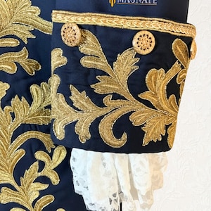 K&ouml;nnte beinhalten: Nahaufnahme eines marineblauen Jacken&auml;rmels mit aufwendiger goldener Stickerei und dekorativen goldenen Kn&ouml;pfen. Die Manschette hat eine wei&szlig;e Spitzenborte. Das Logo "MAGNATE" ist sichtbar. Dieses Kleidungsst&uuml;ck ist wahrscheinlich Teil eines historischen Kost&uuml;ms.