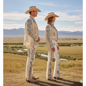 May include: Two people wearing matching cream-colored cowboy outfits with intricate blue embroidery and fringe. They are wearing cowboy hats and boots, standing in a field with a river and mountains in the background. The outfits are detailed and eye-catching.