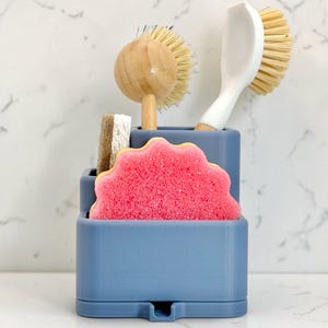 May include: A blue tiered kitchen sink organiser holding a round bamboo dish brush, a white handled scrub brush, a rectangular beige sponge, and a scalloped pink sponge. The organiser features multiple compartments for storing cleaning tools.