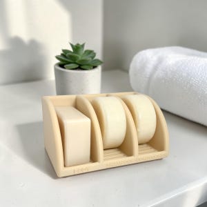 May include: A beige silicone organizer tray with three distinct compartments, each holding a bar of soap. The base features embossed text labels reading BODY, SHAMPOO, and CONDITIONER, with ribbed surfaces inside each section to allow for drainage.
