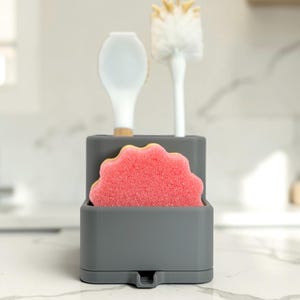 May include: A dark grey rectangular kitchen sink organiser with two compartments. The front section holds a scalloped pink sponge, while the back section contains a white silicone spatula with a wooden handle and a white bottle cleaning brush.