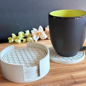 May include: A set of light gray circular coasters featuring a raised hexagonal pattern, stacked within a matching cylindrical holder. One coaster sits outside the holder, supporting a matte black mug with a bright yellow interior lining.