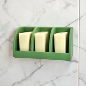 May include: A matte olive green wall-mounted soap holder featuring three distinct vertical compartments. Each section contains a single rectangular bar of off-white soap, neatly organized and held upright within the structured green frame.
