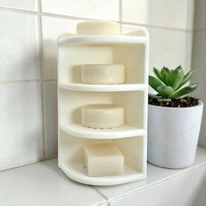 May include: A white, multi-tiered corner storage unit featuring four curved shelves. Three round, off-white soap bars are placed on the top three shelves, while a single rectangular, off-white soap bar sits on the bottom shelf of the organizer.