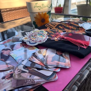 May include: A collection of colorful stickers, photos, and a black t-shirt with the text "The" on it. The items are arranged on a brown wicker surface with a pink surface underneath.