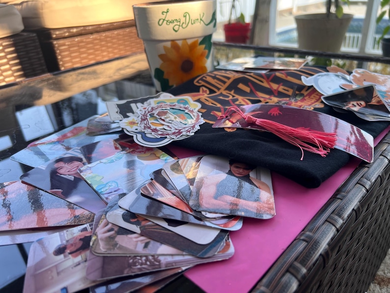 May include: A collection of colorful stickers, photos, and a black t-shirt with the text "The" on it. The items are arranged on a brown wicker surface with a pink surface underneath.