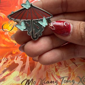 May include: A metal pin depicting a red umbrella with three light blue butterflies. The umbrella has a dark grey handle and trim. The pin is held in a hand, with red glitter nail polish and gold nail art.