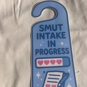 May include: A light blue door hanger with the text "SMUT INTAKE IN PROGRESS". The design includes a progress bar with hearts and a notepad with hearts and a pen. Decorative stars are also included.