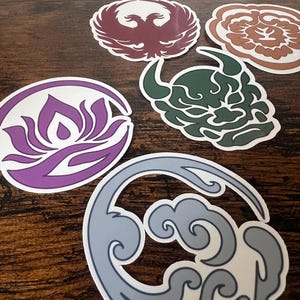 May include: A collection of die-cut stickers featuring various designs. The stickers include a phoenix, a floral pattern, a demon face, a lotus flower, and a wave pattern. The stickers are in shades of purple, brown, green, and blue, with white borders.