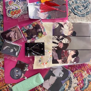 May include: A collection of various items including stickers, bookmarks, a tote bag, a necklace, a small tin, and a plastic bag. The items are all on a pink surface. The tote bag features a black and white illustration of several men.