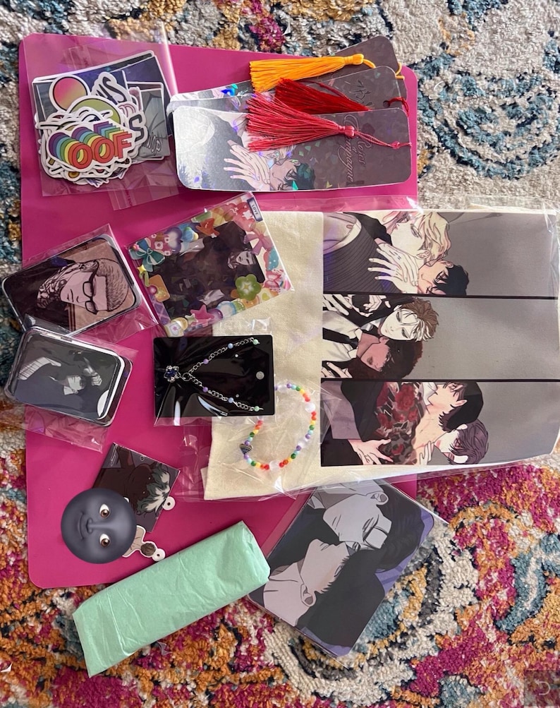 May include: A collection of various items including stickers, bookmarks, a tote bag, a necklace, a small tin, and a plastic bag. The items are all on a pink surface. The tote bag features a black and white illustration of several men.