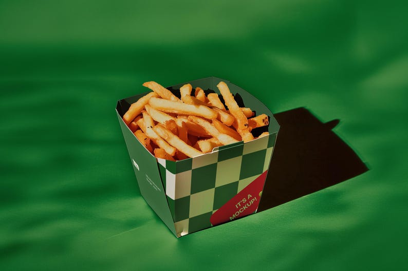 K&ouml;nnte beinhalten: Ein gr&uuml;ner und wei&szlig;er Schachtelkarton, gef&uuml;llt mit goldbraunen Pommes Frites. Auf der Seite des Beh&auml;lters steht der Text "It's a Mockup!"