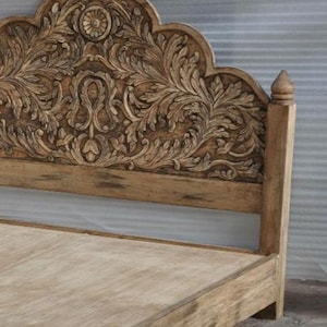 May include: A light brown wooden bed frame featuring an arched headboard with intricate, deep-relief floral and scroll carvings. The headboard is topped with a central circular medallion and features a smooth, flat wooden base frame.