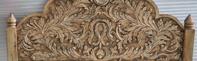 May include: A light brown wooden headboard featuring intricate hand-carved floral and scrolling leaf patterns across the entire surface. The top edge has a curved, arched silhouette with a central decorative motif and pointed finials on each side.