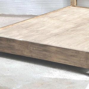May include: A low-profile bed frame constructed from light brown wood with a visible grain pattern. The frame features a rectangular base with four sturdy corner posts and a flat platform surface designed to support a mattress without a box spring.