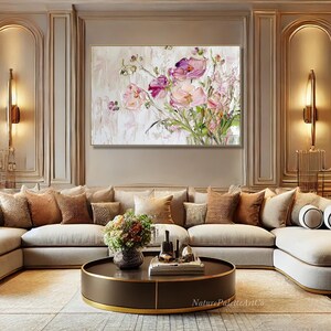May include: A large, abstract painting of pink and purple flowers in a vase. The painting is on a white background and is framed in a gold frame. The painting is hanging on a wall in a living room with a beige sectional sofa and a brown coffee table.